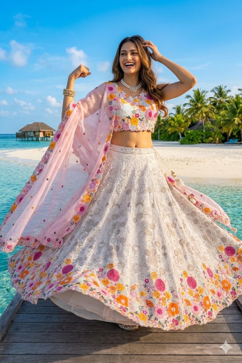 May include: A white and floral embroidered lehenga with a matching cropped top and a sheer pink dupatta. The outfit features colorful floral embroidery and is worn on a wooden dock near the ocean.