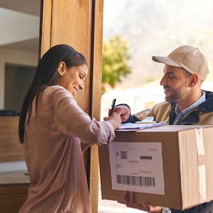 May include: A woman signing for a package delivered by a delivery person. The cardboard box has a shipping label with a barcode and the letter "P". The delivery person is wearing a tan cap and jacket.