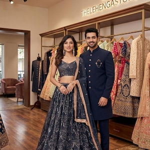 Peut inclure: Un couple pose dans une boutique, présentant des tenues indiennes formelles. La femme porte un lehenga bleu marine avec des détails dorés, tandis que l'homme est en costume bleu marine. L'enseigne du magasin indique "THELEHENGASSTORE".