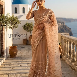 May include: A woman in a beige sari with gold sequin detailing. The sari is paired with a matching blouse. The background includes a blue-domed building, a stone balcony, and a sea view. The text "THE LEHENGA STORE" is visible.