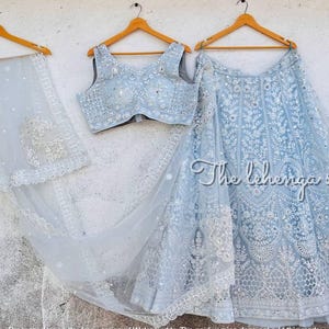 以下が含まれることがあります： 木製のハンガーに掛けられた、ライトブルーの刺繍入りレヘンガセット。スカート、トップス、ドゥパッタが含まれています。セットは複雑な白い刺繍と「The lehenga store」のテキストが特徴です。