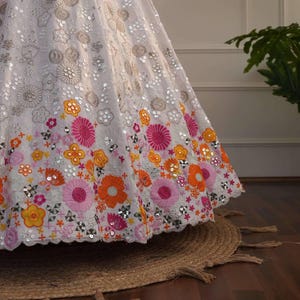 May include: A white, embellished skirt with floral embroidery in shades of pink, orange, and yellow. The skirt features intricate sequin detailing and a scalloped hemline. The skirt is part of a formal wear collection.