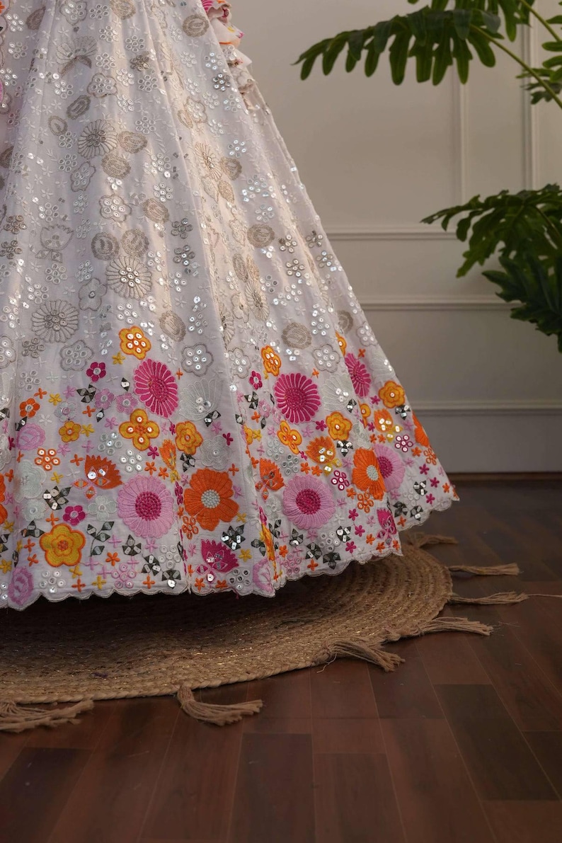 May include: A white, embellished skirt with floral embroidery in shades of pink, orange, and yellow. The skirt features intricate sequin detailing and a scalloped hemline. The skirt is part of a formal wear collection.