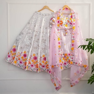 May include: A white skirt and a pink top with a matching scarf are displayed on hangers. The skirt is embellished with silver sequins and colorful floral embroidery in pink, orange, and yellow. The top and scarf feature similar floral designs.