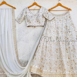May include: A white lehenga with gold embroidery, a matching white embroidered blouse, and a white dupatta. The lehenga has a floral pattern and the blouse has a V-neckline. The dupatta is plain white.