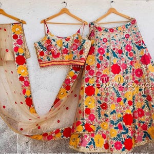 May include: A three-piece floral embroidered Indian outfit. The set includes a crop top, a long skirt, and a sheer scarf. The outfit features red, yellow, and pink flowers with green leaves on a beige background. The outfit is hanging on wooden hangers.