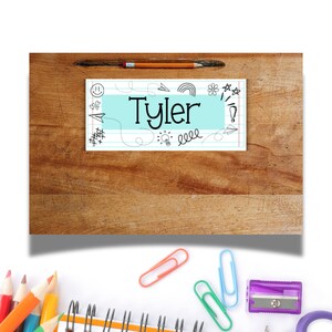 Puede incluir: Una etiqueta de nombre azul y blanca con el nombre "Tyler" escrito en negro. La etiqueta tiene un fondo azul claro con un borde blanco y est&aacute; decorada con varios garabatos, incluyendo un arco&iacute;ris, una bombilla y una cara sonriente.