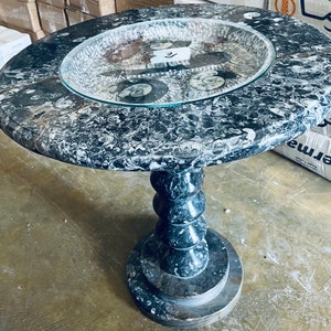 May include: A round, black and white marble table with a glass top. The table top is covered in fossilized ammonites. The table has a pedestal base with a spiral design.
