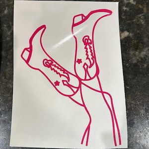 May include: A pink vinyl decal of a pair of cowboy boots with stars on them. The boots are in a crossed position, with one boot pointing up and the other pointing down.