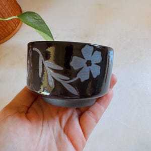 May include: A small, cylindrical ceramic bowl with a glossy black glaze. The exterior features a light gray, etched floral design consisting of a five-petaled flower and a leafy stem. The base of the bowl is unglazed, showing a matte dark gray finish.