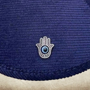 May include: A navy blue corduroy hat with a silver Hamsa hand charm with a blue evil eye in the center.