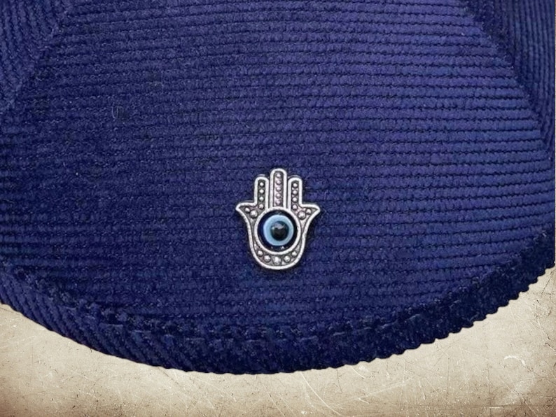 May include: A navy blue corduroy hat with a silver Hamsa hand charm with a blue evil eye in the center.
