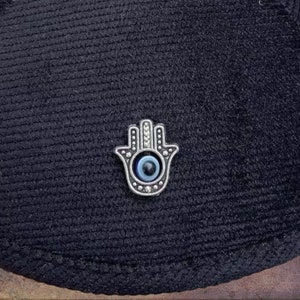 May include: A silver hamsa hand charm with a blue evil eye in the center. The charm is on a black fabric background.