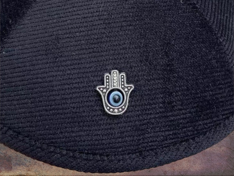 May include: A silver hamsa hand charm with a blue evil eye in the center. The charm is on a black fabric background.