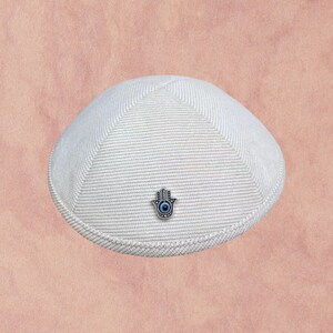 May include: A white knitted kippah with a silver Hamsa hand charm with a blue eye in the center.