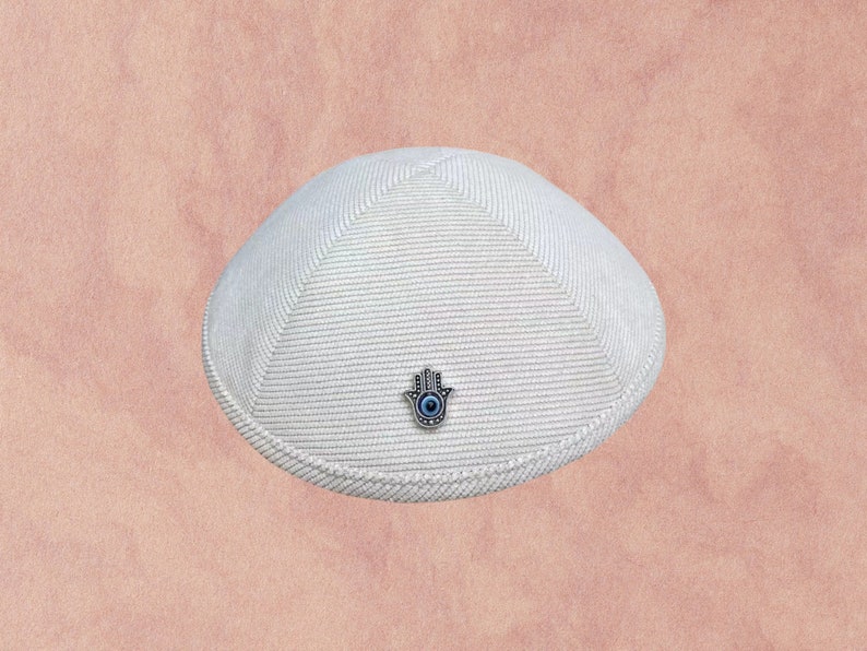 May include: A white knitted kippah with a silver Hamsa hand charm with a blue eye in the center.