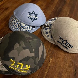 Può includere: Quattro kippah in vari colori e fantasie sono esposte su una superficie di legno. Una è azzurra con una stella di David e la scritta "ISRAEL STRONG". Un'altra è mimetica con testo ebraico e "ISRAEL STRONG". Le altre sono grigie e bianche.