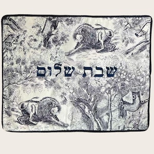 May include: A decorative textile with a black and white toile design featuring lions and trees. The phrase "Shabbat Shalom" is written in blue Hebrew script across the center. The edges are trimmed with a black rope.