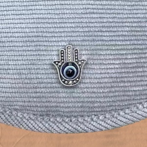 May include: A silver hamsa hand charm with a blue evil eye in the center. The charm is on a gray textured fabric background.