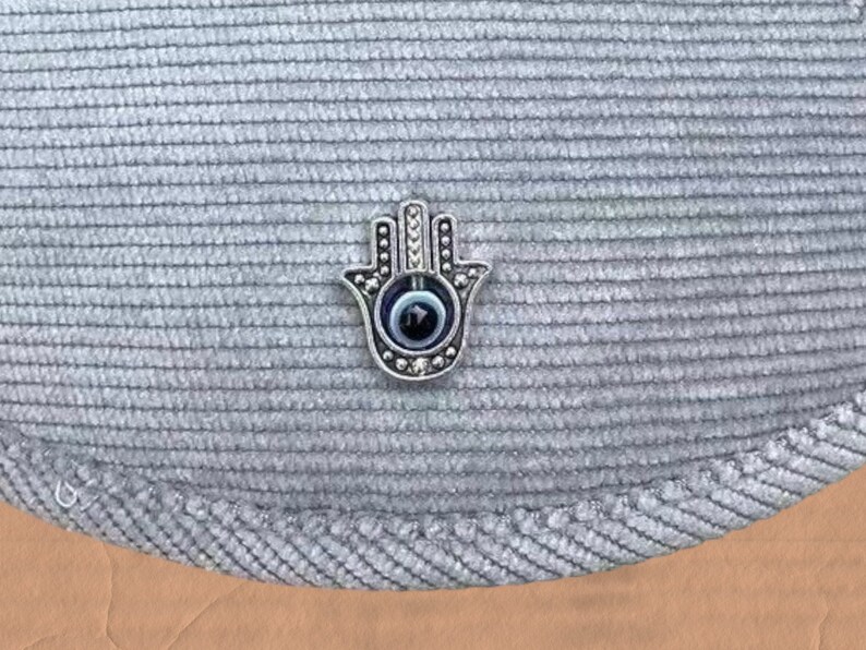 May include: A silver hamsa hand charm with a blue evil eye in the center. The charm is on a gray textured fabric background.
