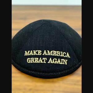 Puede incluir: Una kip&aacute; negra con el texto MAKE AMERICA GREAT AGAIN bordado en hilo dorado. El cubrecabezas tiene una construcci&oacute;n redondeada de varios paneles con una costura circular en la parte superior y un borde reforzado a lo largo de la base.