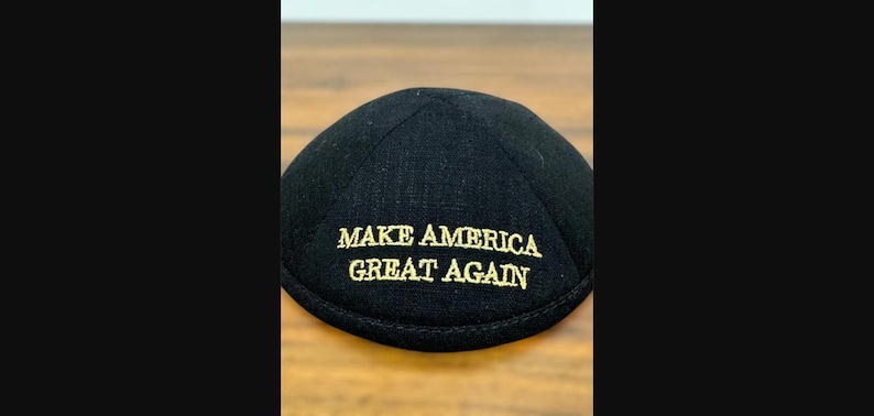 Puede incluir: Una kip&aacute; negra con el texto MAKE AMERICA GREAT AGAIN bordado en hilo dorado. El cubrecabezas tiene una construcci&oacute;n redondeada de varios paneles con una costura circular en la parte superior y un borde reforzado a lo largo de la base.