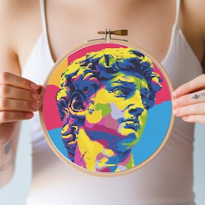 Peut inclure: Un motif de point de croix coloré d'un visage d'homme dans un cerceau de broderie en bois. Le motif est de style pop art avec des couleurs vives et des lignes audacieuses. Le motif est disponible en téléchargement PDF.