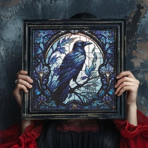 Pode incluir: Uma mulher segura uma obra de arte em vitral emoldurada que retrata um corvo azul empoleirado em um galho. A obra está sobre um fundo azul escuro com intrincados desenhos florais.