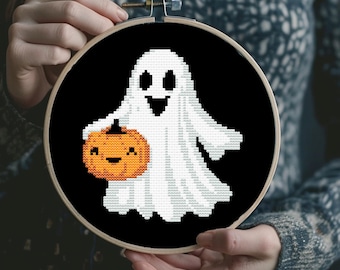 Puede incluir: Un fantasma blanco sosteniendo una pequeña calabaza naranja con una cara sonriente, bordado en punto de cruz sobre tela negra en un aro de bordado redondo.