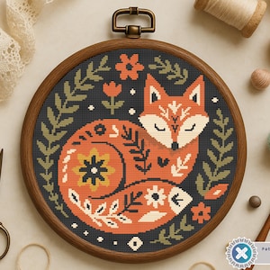 May include: A cross-stitch embroidery hoop featuring a stylized orange fox with floral and leaf accents on a black background. The design is set within a wooden hoop, with a brass hanger. Craft supplies, including scissors and thread, are visible.