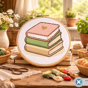 May include: A cross-stitch embroidery hoop featuring a stack of three books in pastel colors: pink, green, and tan. The hoop is set on a wooden table with other craft supplies, including yarn, scissors, and a basket of flowers.
