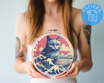 May include: Cross stitch pattern featuring a blue cat sitting in front of a wave with a pink and orange sunset in the background. The pattern is in a wooden embroidery hoop.