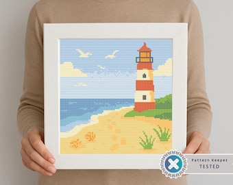 Puede incluir: Un bordado enmarcado que representa una escena costera con un faro. El faro es rojo y blanco, sobre una playa de arena con hierba verde. El cielo es azul con nubes y gaviotas. El marco es blanco.