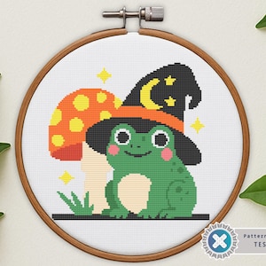 May include: Cross-stitch design of a green frog wearing a witch hat with a crescent moon and stars, and a mushroom. The design is in a wooden embroidery hoop. The frog is smiling. The mushroom is orange with yellow spots.