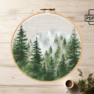May include: A cross-stitch pattern of a forest scene with tall green trees and a white, foggy background. The pattern is displayed in a round wooden embroidery hoop.