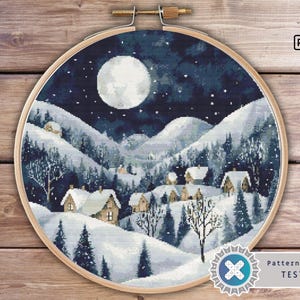 Puede incluir: Un patrón de punto de cruz que representa una escena invernal con luna llena, colinas cubiertas de nieve y un pequeño pueblo de casas. El patrón está en un aro redondo y está etiquetado como "Pattern Keeper TESTED".
