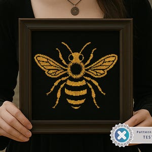 May include: Framed cross-stitch art featuring a golden bee on a black background. The bee is detailed with wings, antennae, and body segments. The artwork is in a dark brown frame. The image includes a "Pattern Keeper TESTED" badge.