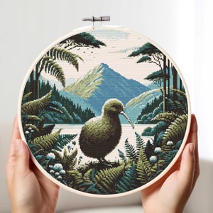 May include: A cross-stitch embroidery hoop depicting a kiwi bird standing in a lush green forest with a mountain range in the background.