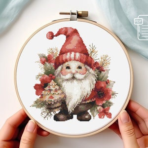 May include: Cross stitch pattern of a red and white gnome holding a small Christmas tree. The gnome is surrounded by red flowers and greenery.