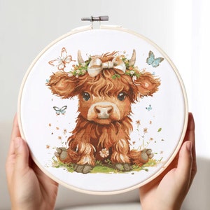 May include: A cross-stitch embroidery hoop featuring a charming brown and white Highland cow wearing a flower crown and a white bow. The cow is sitting on a green patch of grass with butterflies and flowers around it.