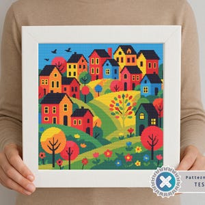 Può includere: Paesaggio a punto croce incorniciato, raffigurante un villaggio colorato con case, alberi e fiori. L'opera utilizza tonalità rosse, gialle, blu e verdi su uno sfondo di cielo blu. La cornice è bianca.