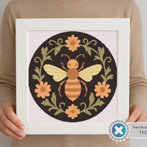 May include: Framed cross-stitch art featuring a bee design. The bee is in the center, surrounded by flowers and leaves. The color palette includes shades of brown, yellow, and orange on a black background. The frame is white.