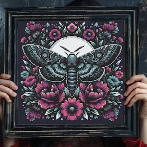 May include: A framed cross-stitch embroidery featuring a dark blue and green moth with a white moon behind it. The moth is surrounded by pink and blue flowers.