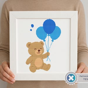 May include: A framed cross-stitch design featuring a brown teddy bear holding three blue balloons. The bear is smiling, and two smaller blue dots are above the balloons. The artwork is in a white frame.