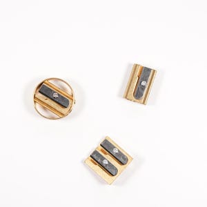 May include: Three gold pencil sharpeners on a white background. One sharpener is round, one is rectangular, and one is square.