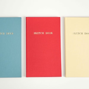 Könnte beinhalten: Drei Skizzenbücher, eines blau, eines rot und eines gelb, jedes mit dem Schriftzug "Sketch Book" in Gold auf dem Einband.