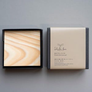 May include: A wooden block memo pad in a black box, with a light brown and beige color scheme. The open box reveals the wood grain pattern. The closed box has text and a logo. The product is on a gray background.