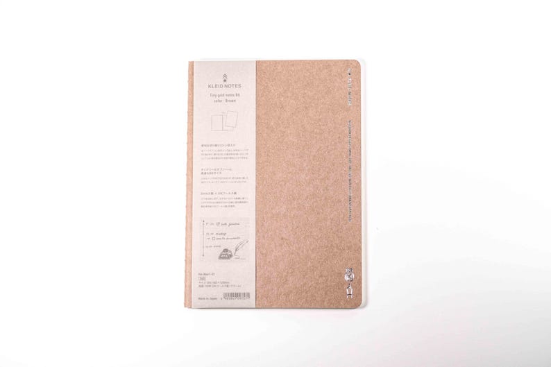 K&ouml;nnte beinhalten: Ein beigefarbenes Notizbuch mit einem Papieretikett auf der linken Seite. Das Etikett enth&auml;lt Text und Illustrationen auf Japanisch und Englisch. Das Notizbuch ist geschlossen und hat ein minimalistisches Design. Das Notizbuch befindet sich auf wei&szlig;em Hintergrund.