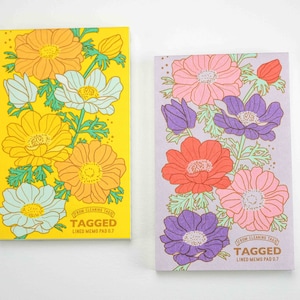 May include: Two lined memo pads with floral designs. The left pad has a yellow background with orange, white, and light blue flowers. The right pad has a light purple background with pink, red, and purple flowers. Both pads have the text "TAGGED" and "LINED MEMO PAD 0.7" printed on them.