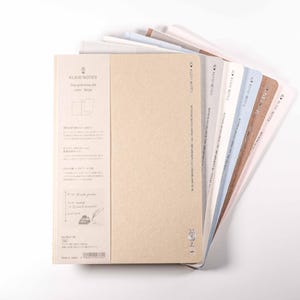 K&ouml;nnte beinhalten: Eine Auswahl an Notizb&uuml;chern mit verschiedenfarbigen Einb&auml;nden, darunter Beige, Wei&szlig;, Blau und Braun, f&auml;cherf&ouml;rmig auf wei&szlig;em Hintergrund angeordnet. Das vordere Notizbuch hat ein Etikett mit Text und Illustrationen. Geeignet zum Schreiben oder Skizzieren.